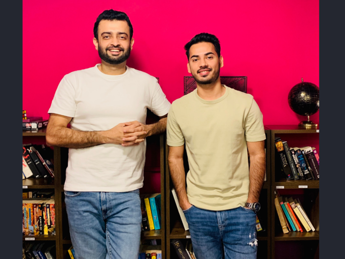 Two smiling individuals stand in front of a vibrant pink wall, leaning against a bookshelf filled with books. Both wear casual jeans and t-shirts, creating a relaxed, friendly atmosphere.