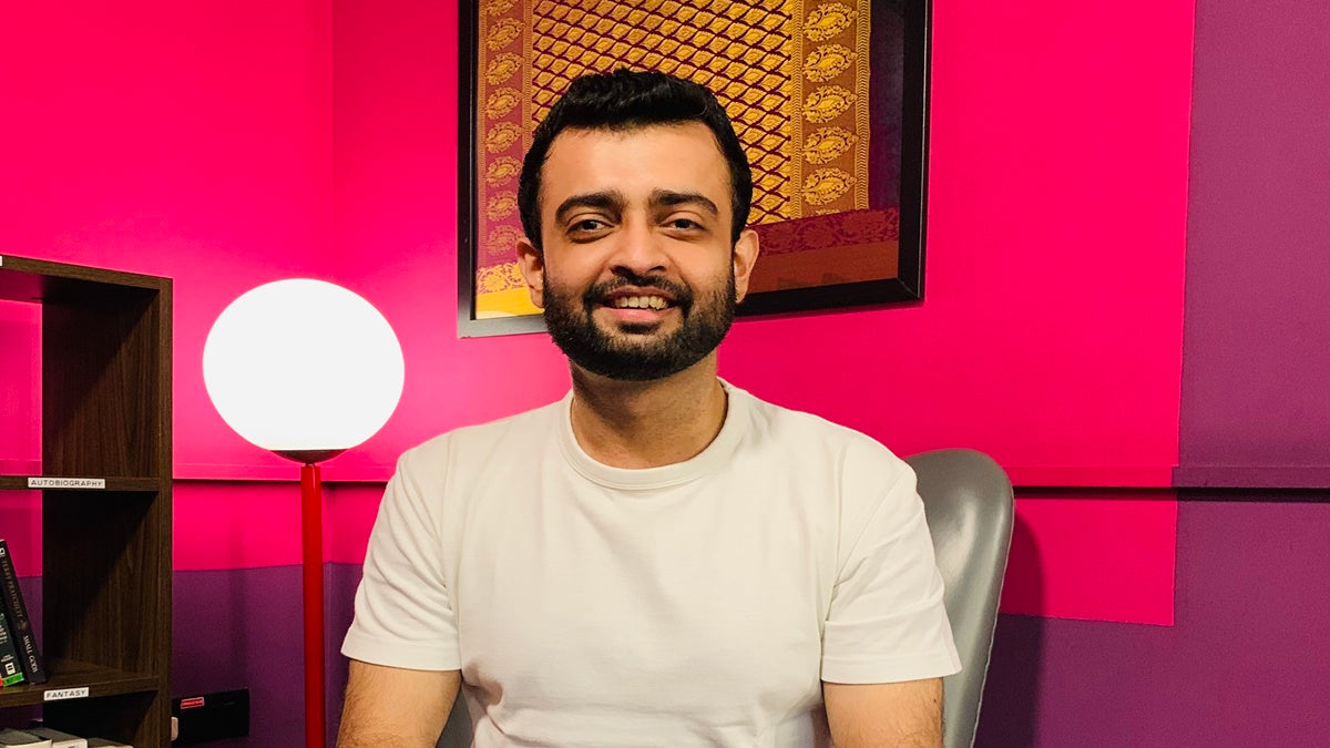 A bearded man sits in front of a vibrant pink wall, creating a striking contrast in the image